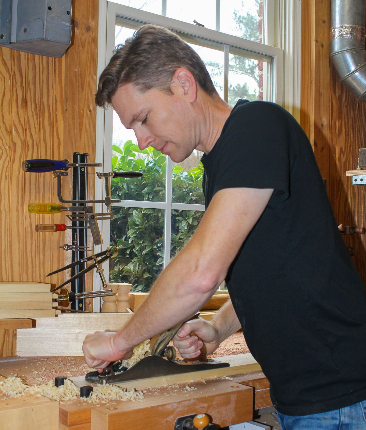 Erin Watson smoothing a board with a hand plane at the workbench, hand tools and a shop window behind him